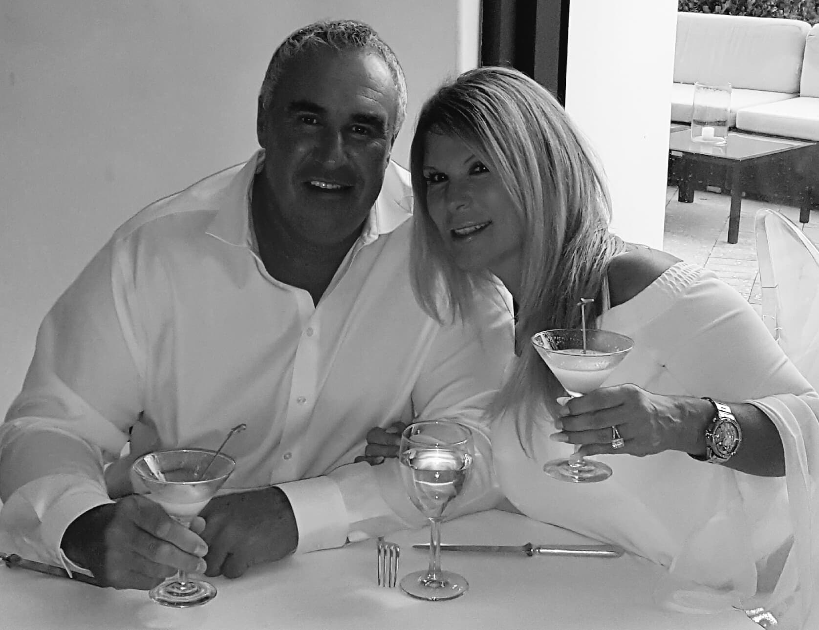 Smiling couple in white holding martini glasses at a table, black and white.
