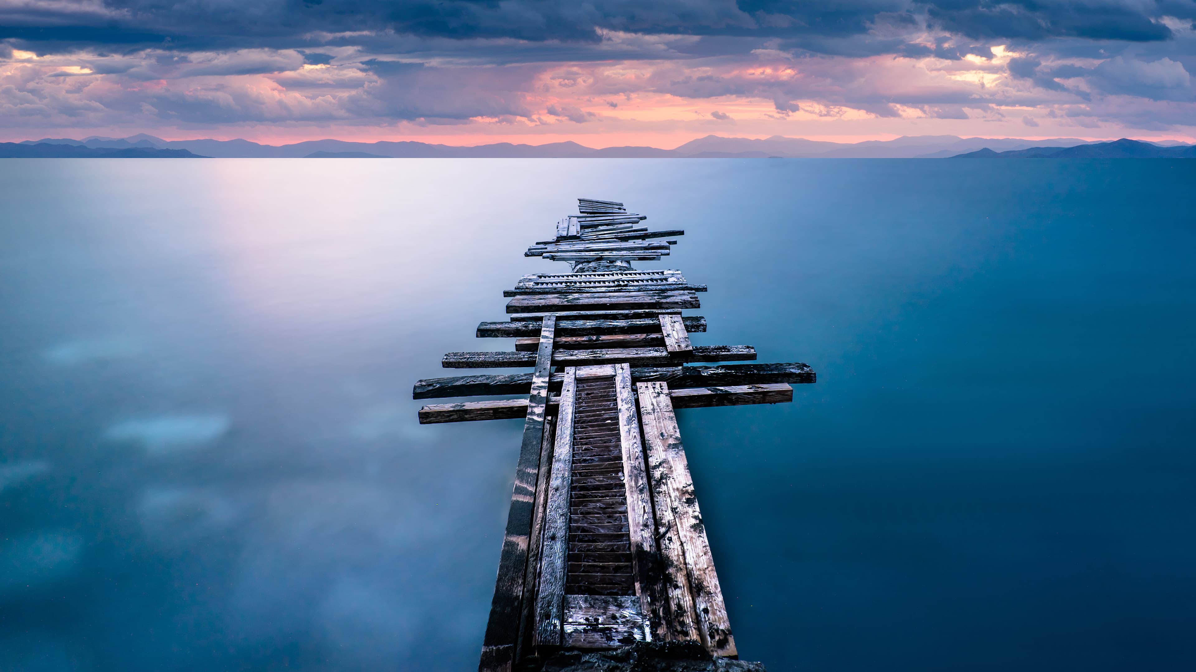 Dilapidated wooden pier stretching into calm blue water under a dramatic sunset sky with mountains.